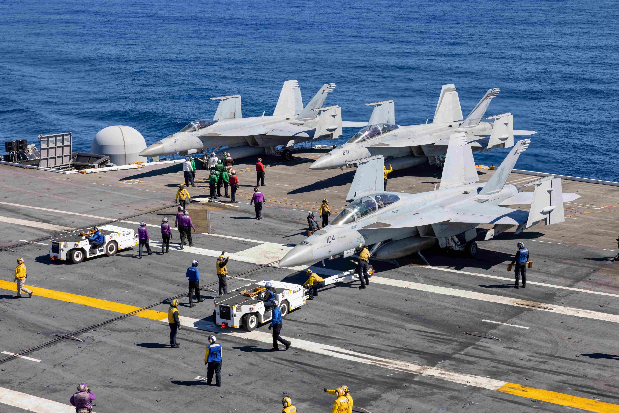 An image of three jets on the flight deck of the USS Nimitz, with the crew bustling