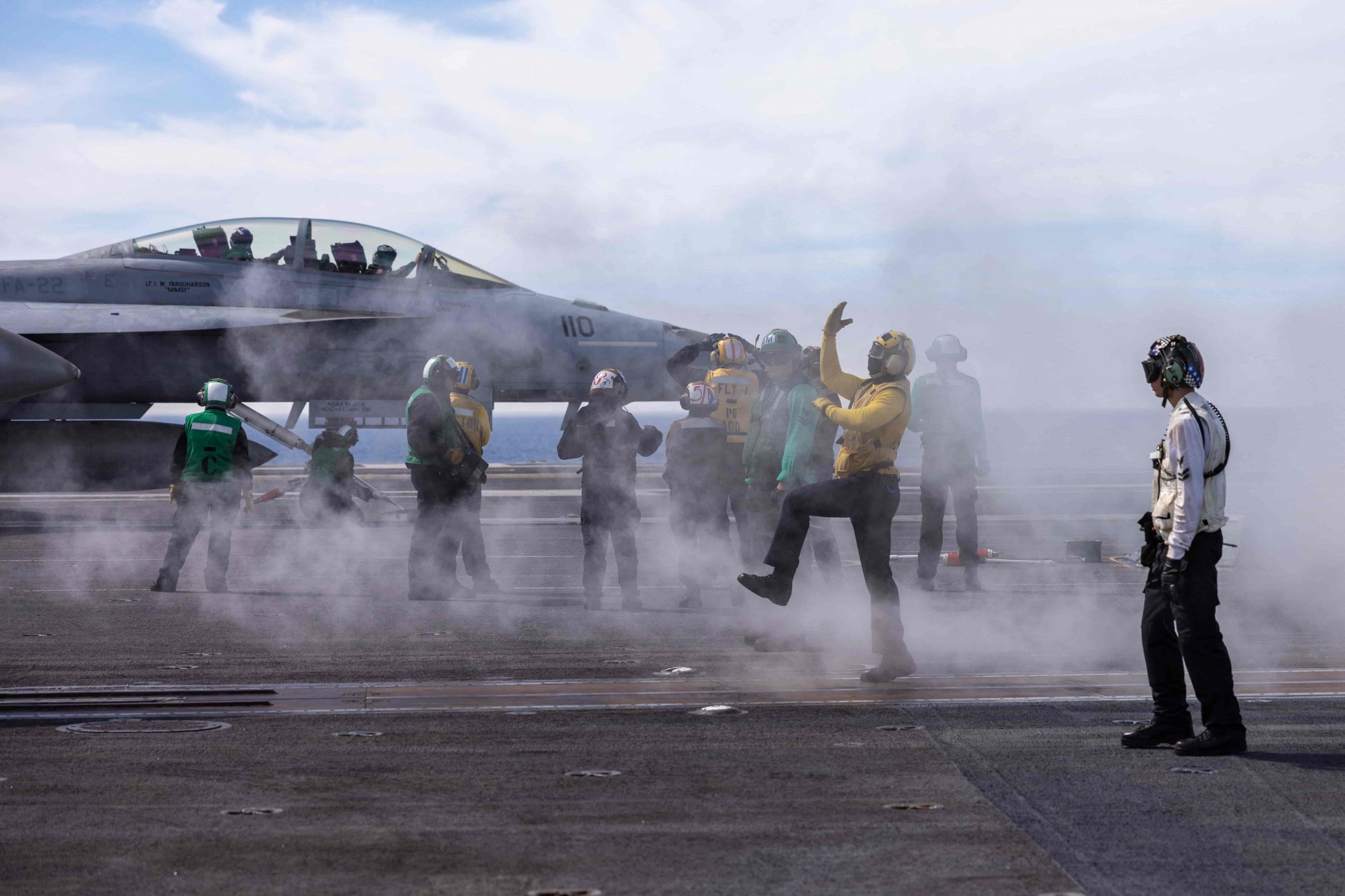 a bunch of flight deck crewmembers standing by a jet, with smoke billowing around them