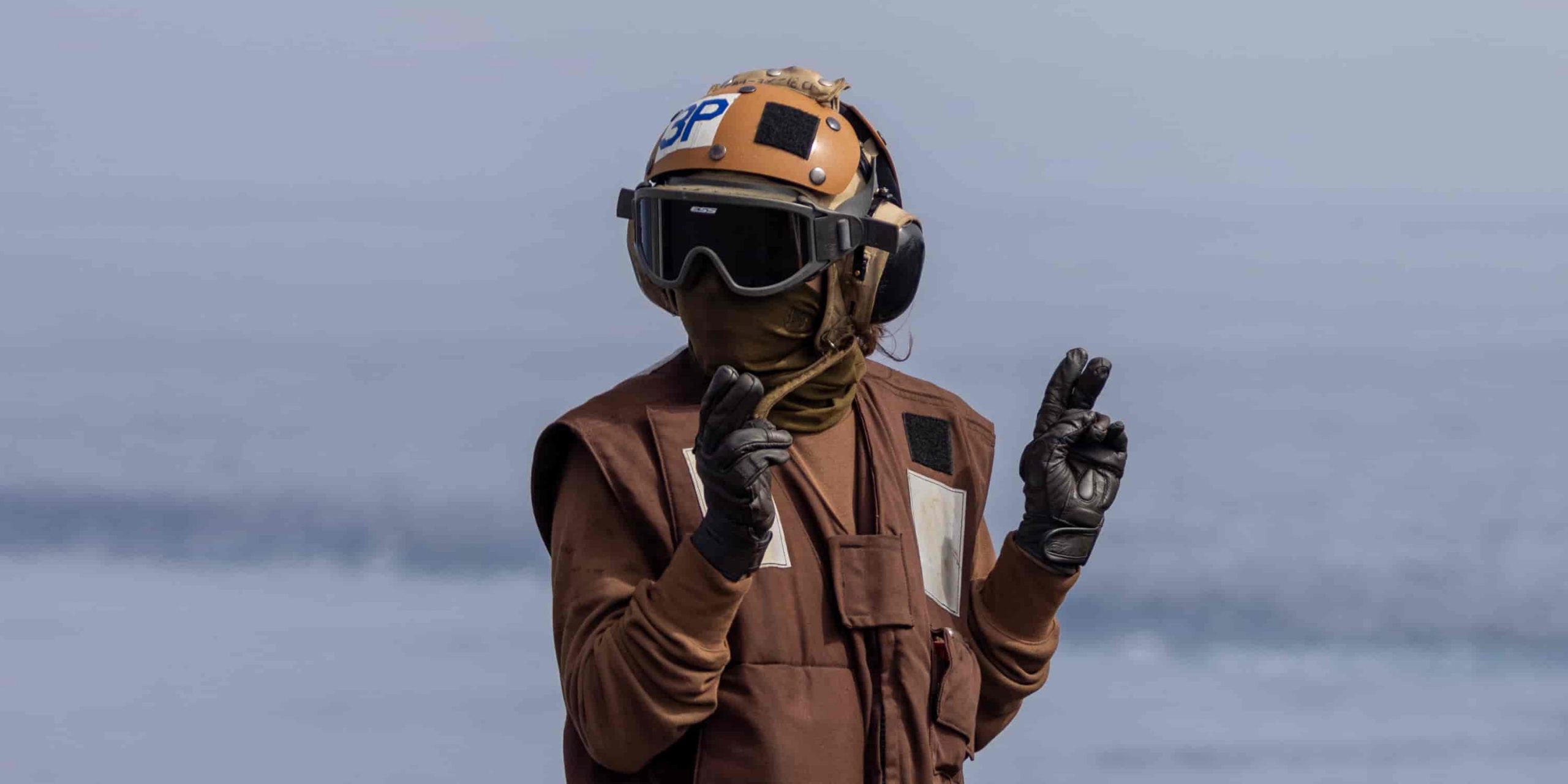 a man on the flight deck stands in brown with his hands up at his sides, holding up two fingers on each hand.