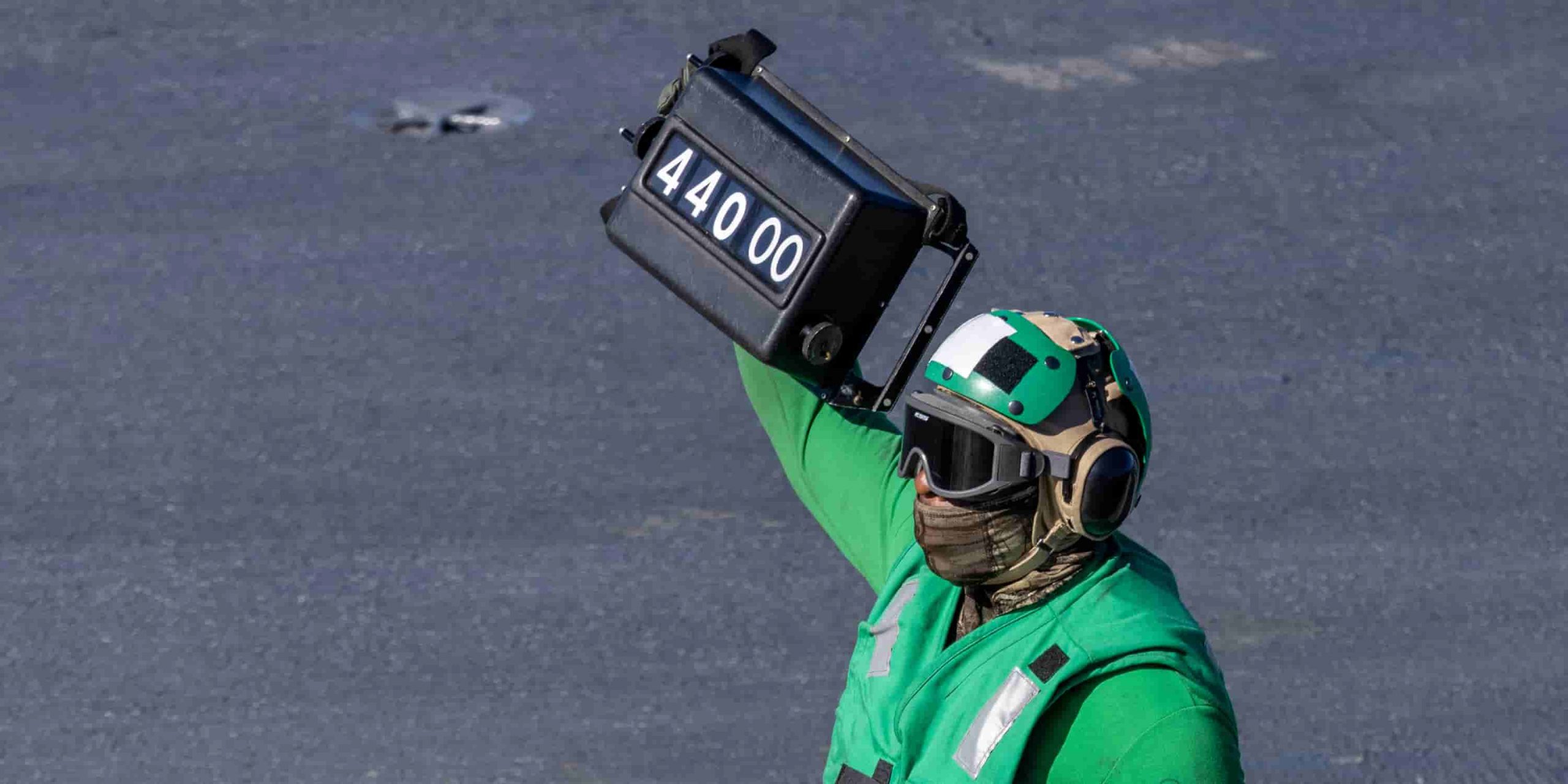 a flight deck officer in green stands hold a box, displaying the number 44000, on his shoulder