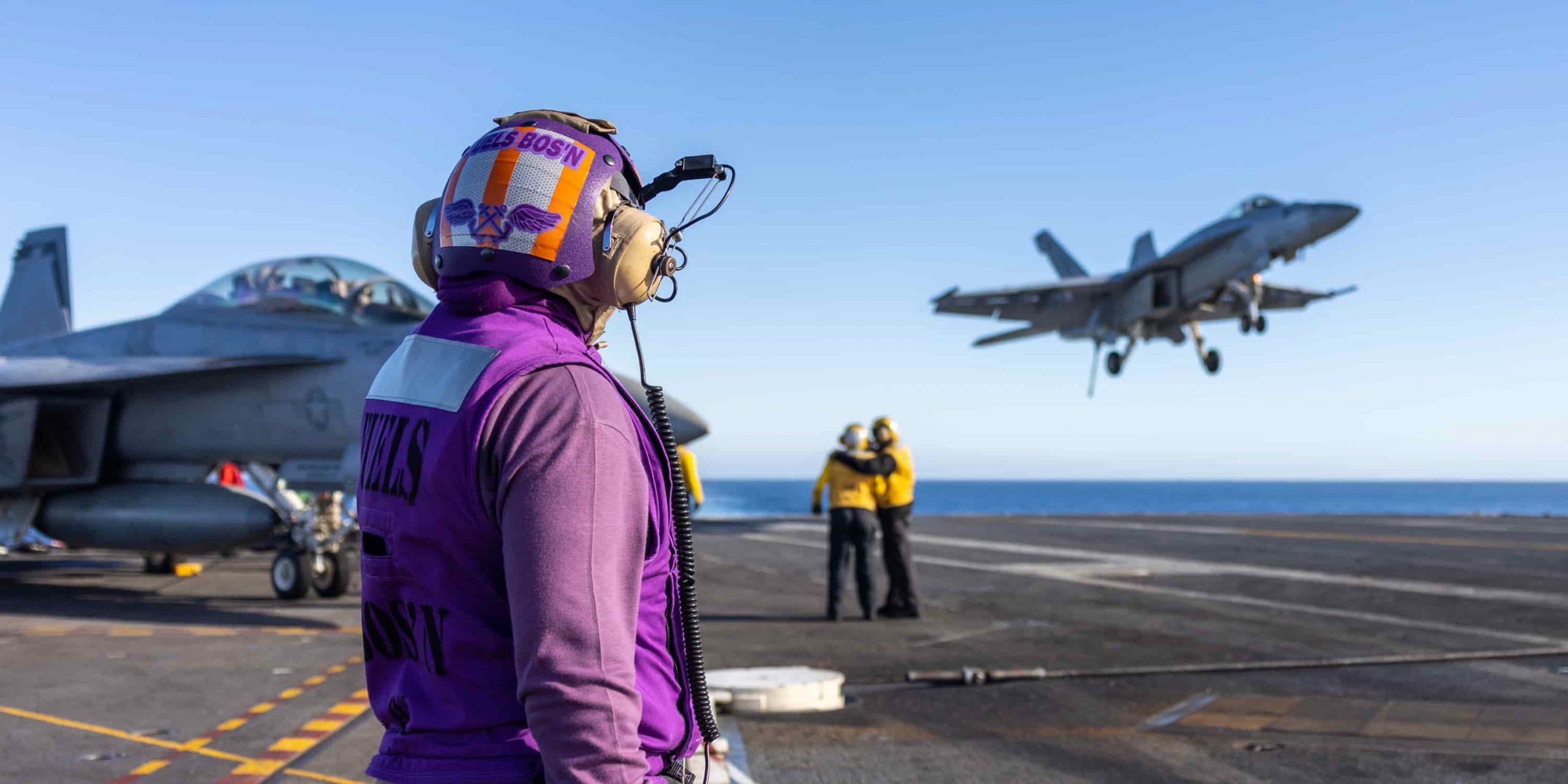 a flight deck officer in purple watches as a jet comes in for landing