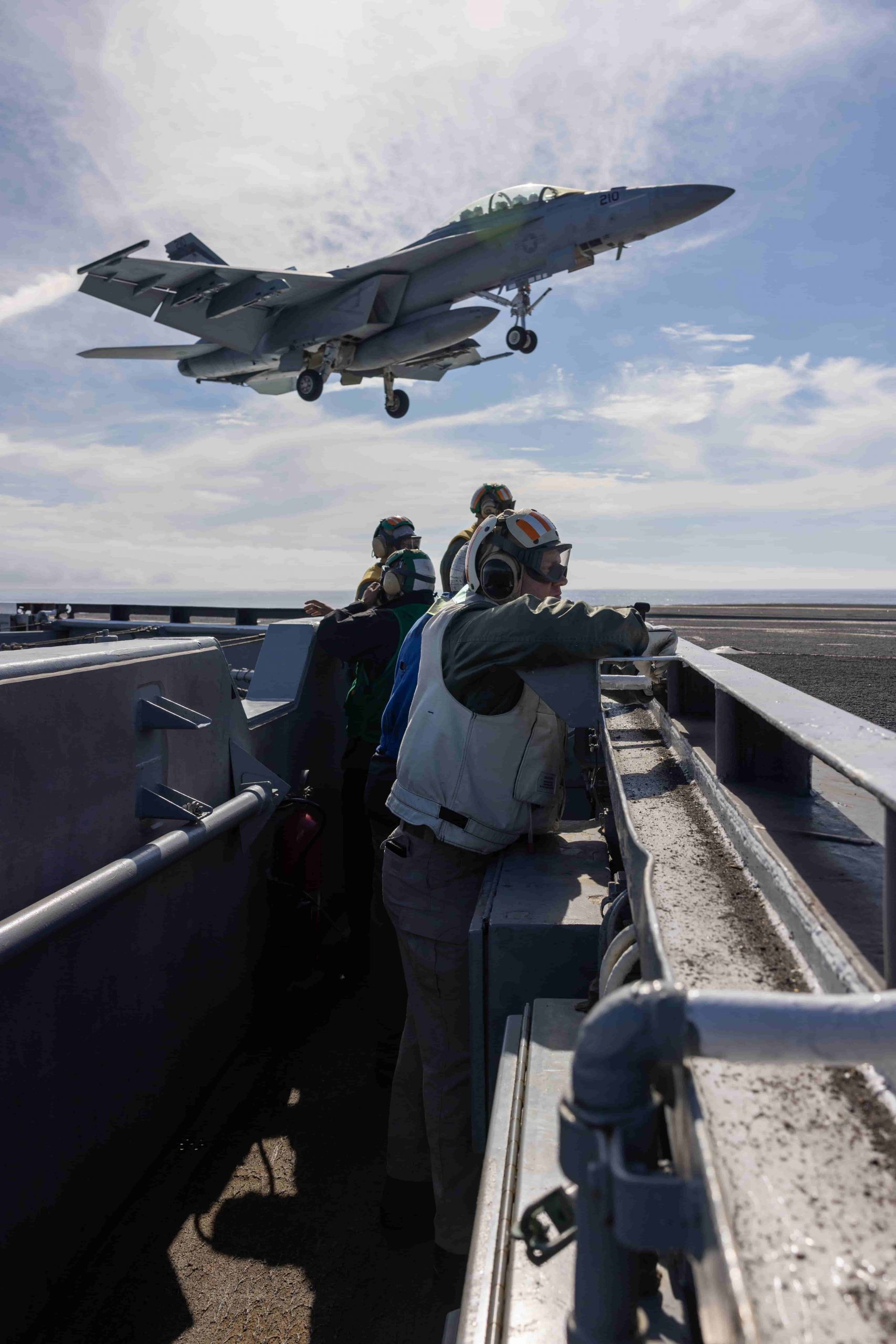 several crew members stand in a "trench" as a jet blasts by overhead