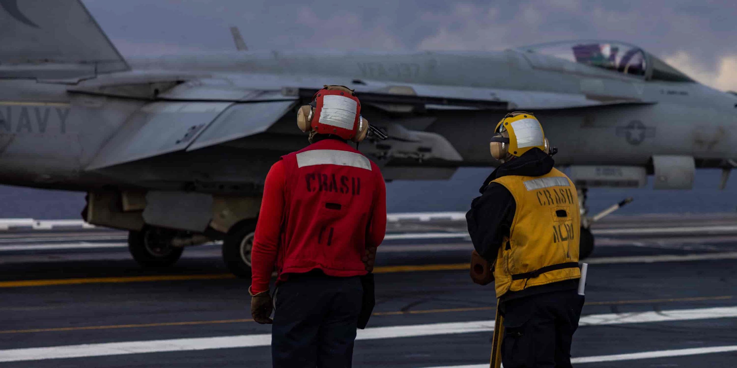 a flight deck officer in red stands next to his colleague in yellow as they chat while facing a jet.